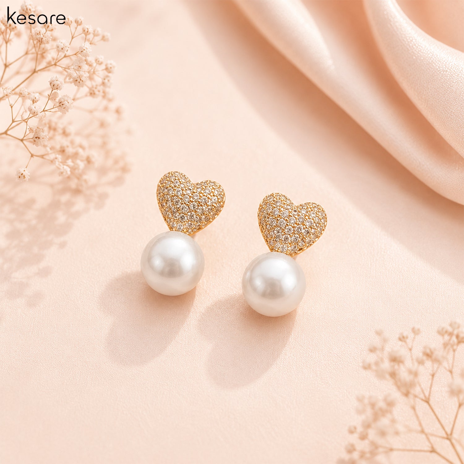A pair of gold heart stud earrings with sparkling pavé tops and round white pearl drops, displayed on a soft peach background with decorative dried flowers and draped satin.