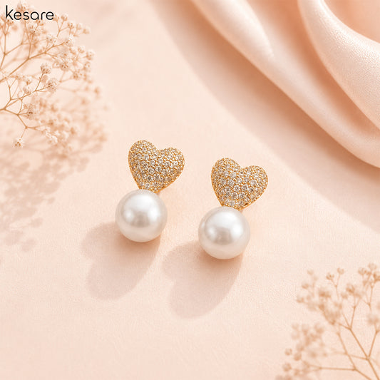 A pair of gold heart stud earrings with sparkling pavé tops and round white pearl drops, displayed on a soft peach background with decorative dried flowers and draped satin.