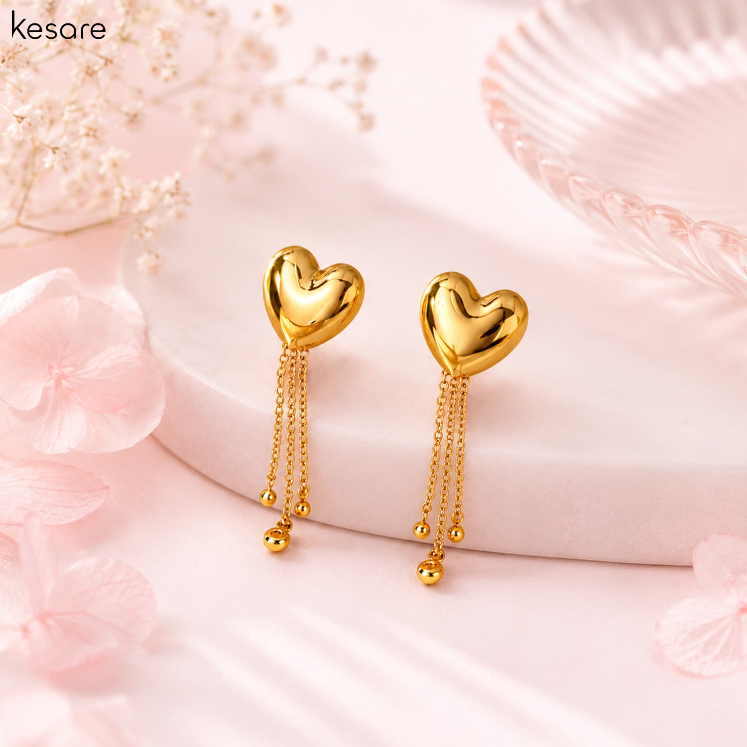 A pair of gold heart-shaped dangle earrings with three hanging chains and small bead drops, displayed on a soft pink surface with decorative flowers and a glass dish. The brand name 'Kesare' is visible in the top left corner.