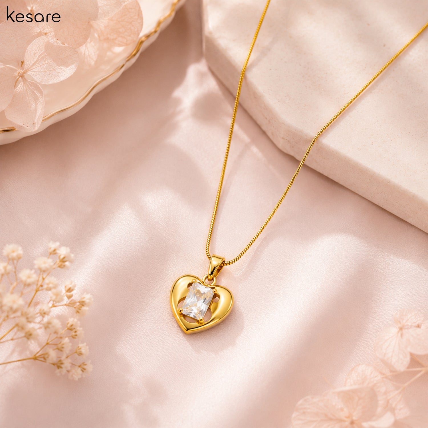 A gold heart pendant necklace with a clear rectangular gemstone, displayed on a soft pink fabric background with decorative dried flowers and a stone prop. The brand name 'Kesare' is visible in the top left corner.