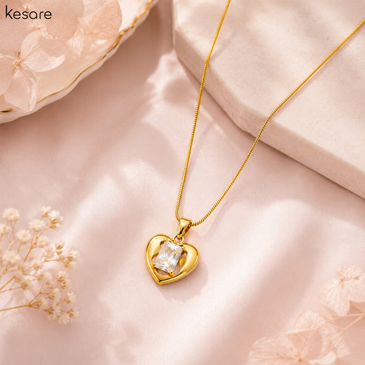 A gold heart pendant necklace with a clear rectangular gemstone, displayed on a soft pink fabric background with decorative dried flowers and a stone prop. The brand name 'Kesare' is visible in the top left corner.