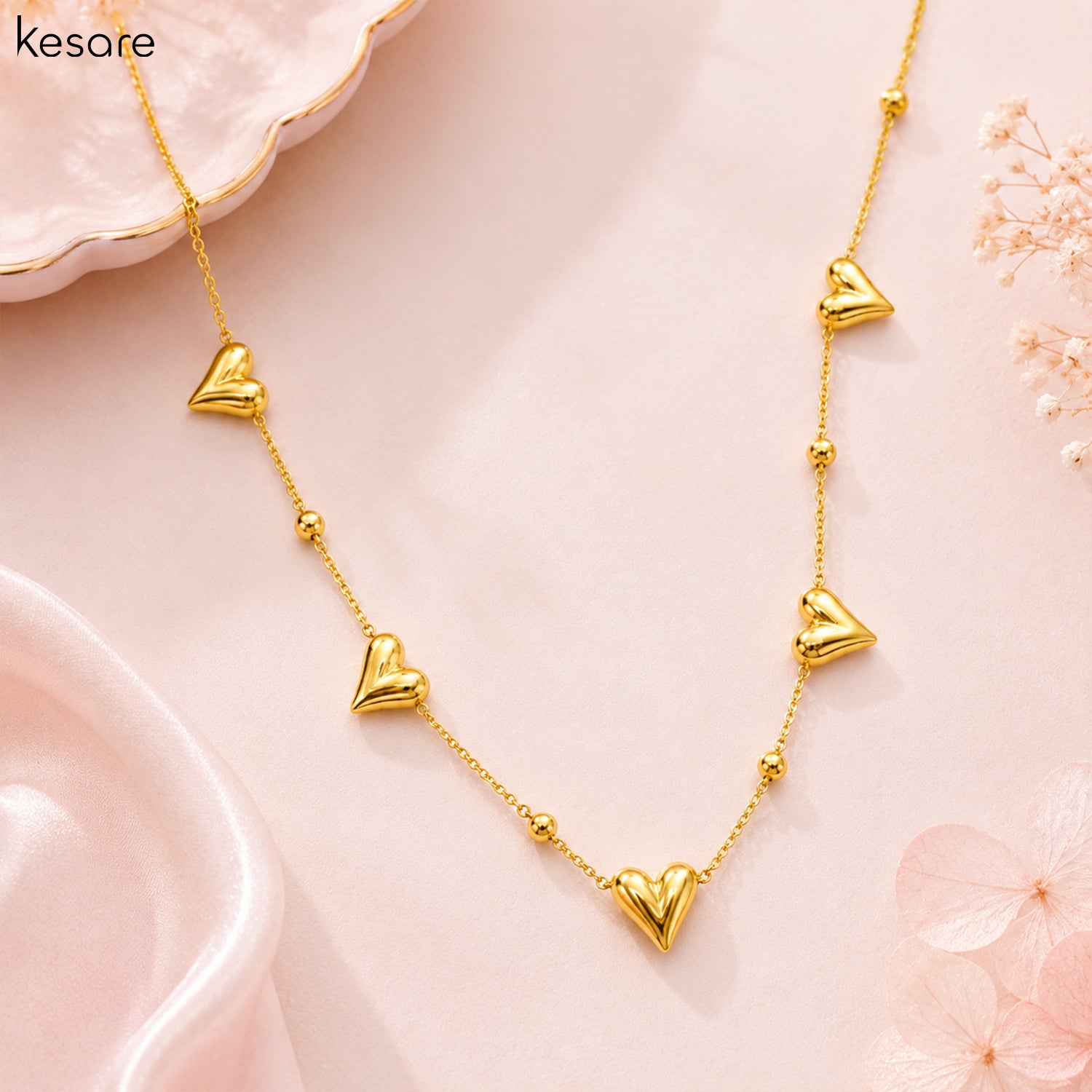 A gold heart necklace with five dangling heart charms and small gold beads on a delicate chain, displayed on a soft pink background with decorative pink petals and a white dish.