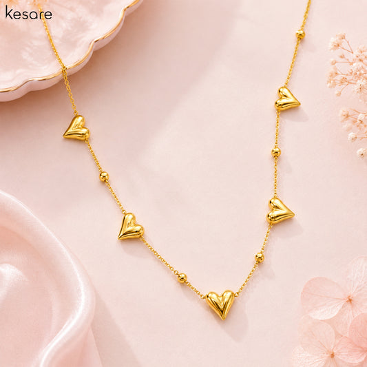 A gold heart necklace with five dangling heart charms and small gold beads on a delicate chain, displayed on a soft pink background with decorative pink petals and a white dish.