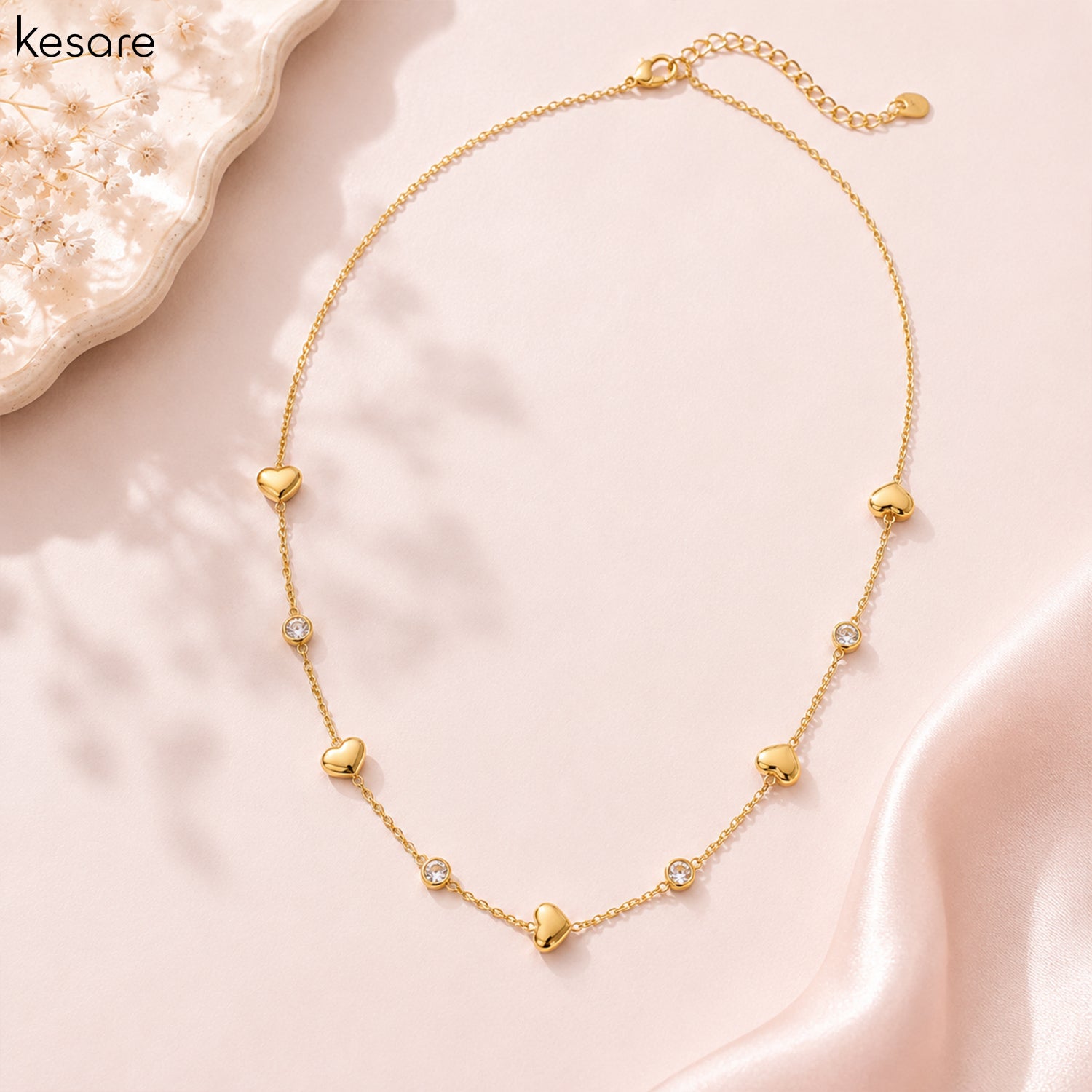 A delicate gold chain necklace with multiple small heart charms and a few clear rhinestone accents, displayed on a soft pink satin fabric background with a decorative crystal plate in the corner. The brand name 'Kesare' is visible in the top left.