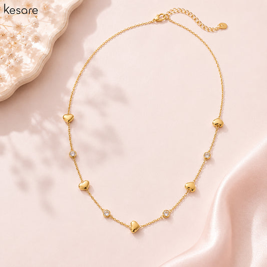 A delicate gold chain necklace with multiple small heart charms and a few clear rhinestone accents, displayed on a soft pink satin fabric background with a decorative crystal plate in the corner. The brand name 'Kesare' is visible in the top left.