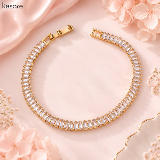 A gold-plated tennis bracelet with clear rectangular baguette crystals, arranged in a circular shape on a soft pink satin background with decorative pink flowers. The brand name 'Kesare' is visible in the top left corner.