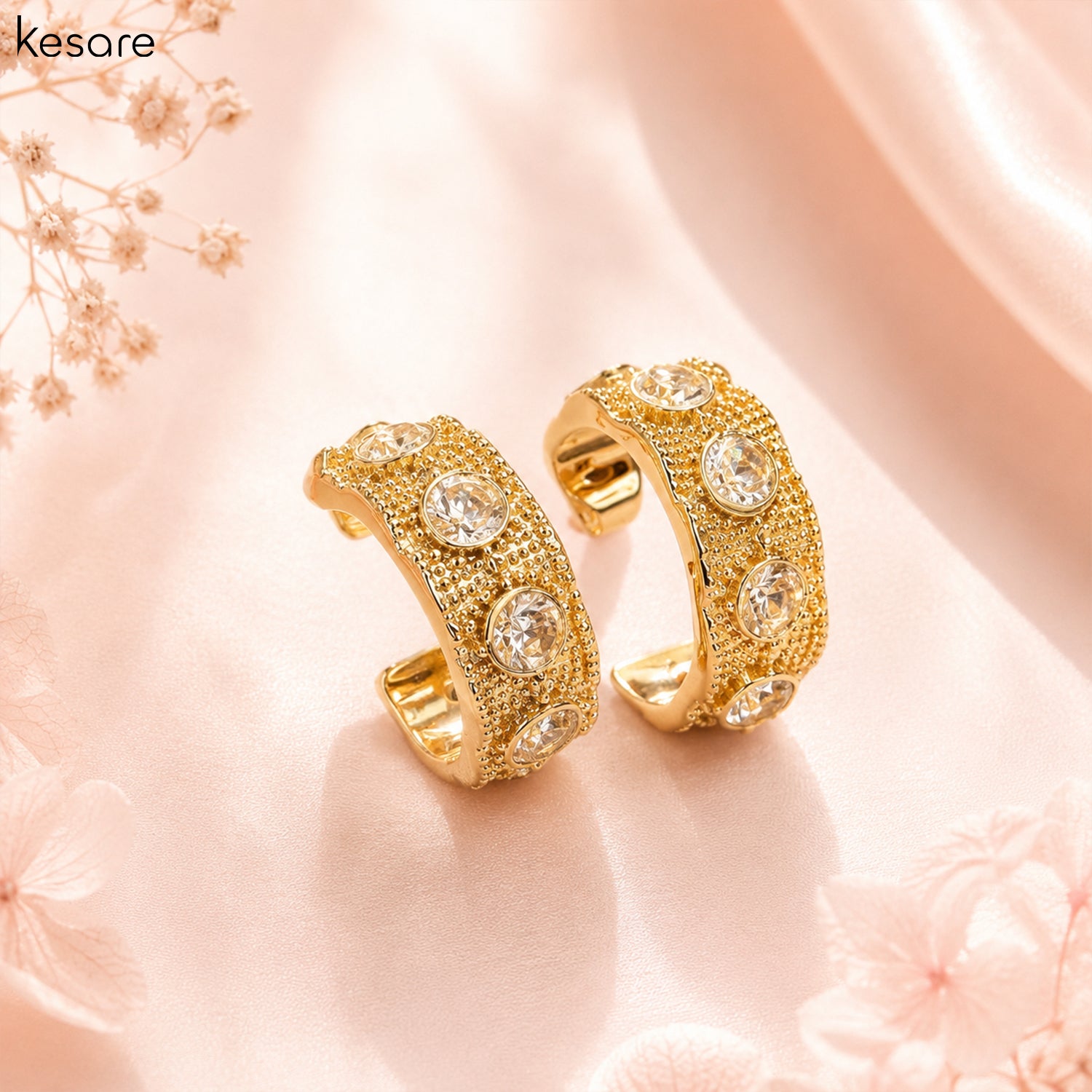 A pair of gold huggie hoop earrings adorned with clear round crystals and textured pavé detailing, displayed on a soft pink satin fabric background with floral props. The brand name 'kesare' is visible in the top left corner.