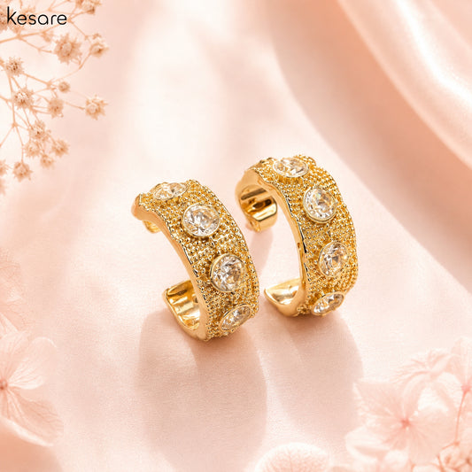 A pair of gold huggie hoop earrings adorned with clear round crystals and textured pavé detailing, displayed on a soft pink satin fabric background with floral props. The brand name 'kesare' is visible in the top left corner.