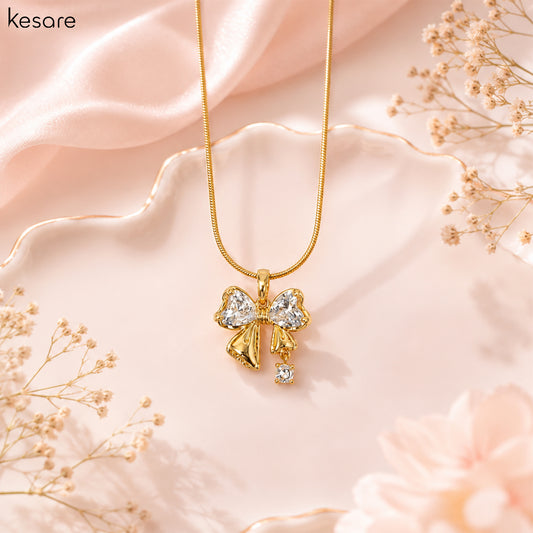 A gold necklace with a bow-shaped pendant adorned with clear crystals, displayed on a soft pink satin background with dried floral decorations. The brand name 'Kesare' is visible in the top left corner.