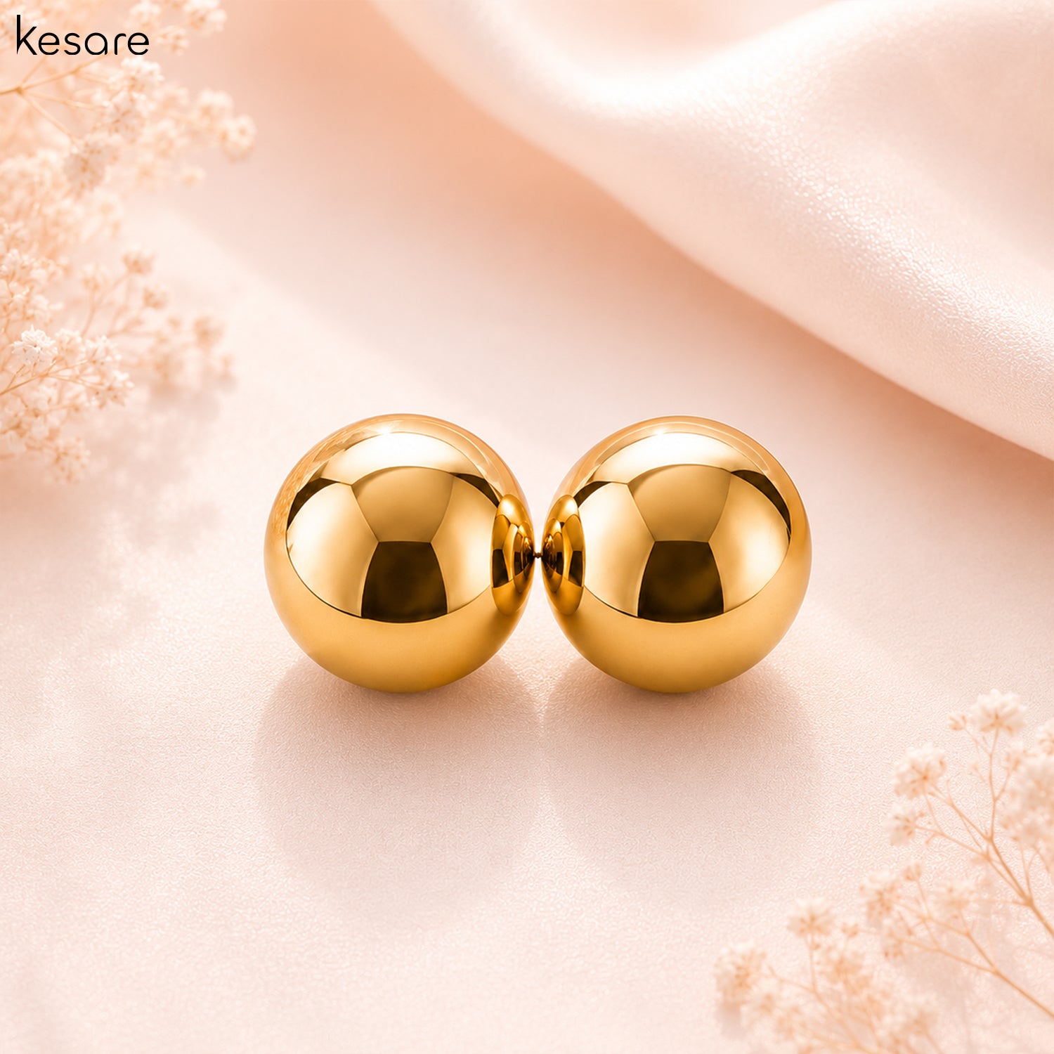 A pair of shiny gold spherical stud earrings displayed on a soft blush pink surface with decorative dried flowers and a draped satin fabric in the background. The brand name 'Kesare' is visible in the top left corner.