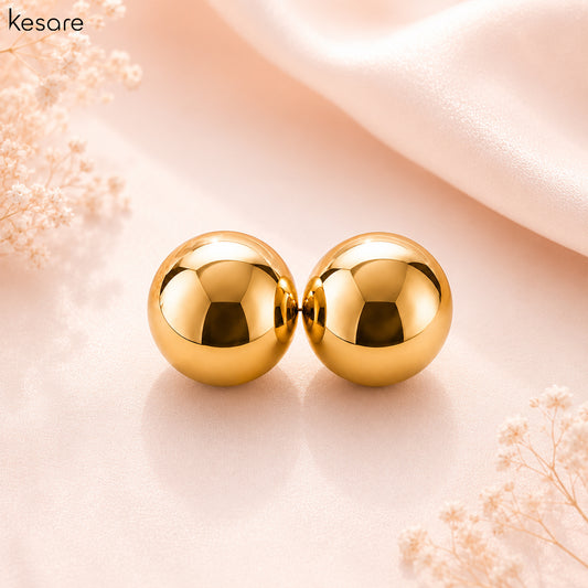 A pair of shiny gold spherical stud earrings displayed on a soft blush pink surface with decorative dried flowers and a draped satin fabric in the background. The brand name 'Kesare' is visible in the top left corner.