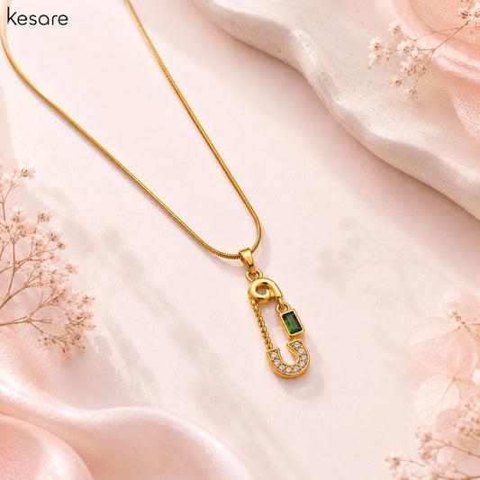 A gold safety-pin style pendant necklace with a green rectangular stone and small clear stones, displayed on a soft pink satin background with dried floral props. The brand name 'Kesare' appears in the top left corner.