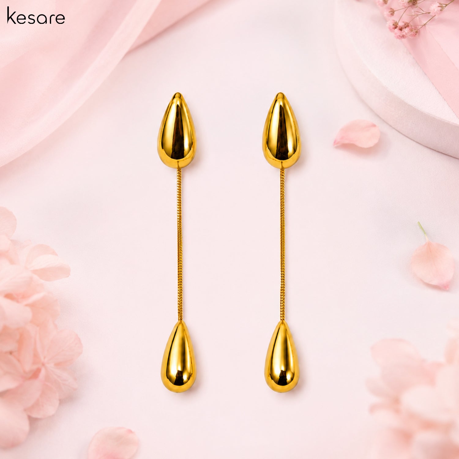 A pair of gold-toned elongated earrings with teardrop-shaped ends and a textured central rod, displayed on a soft pink background with decorative flowers and fabric.