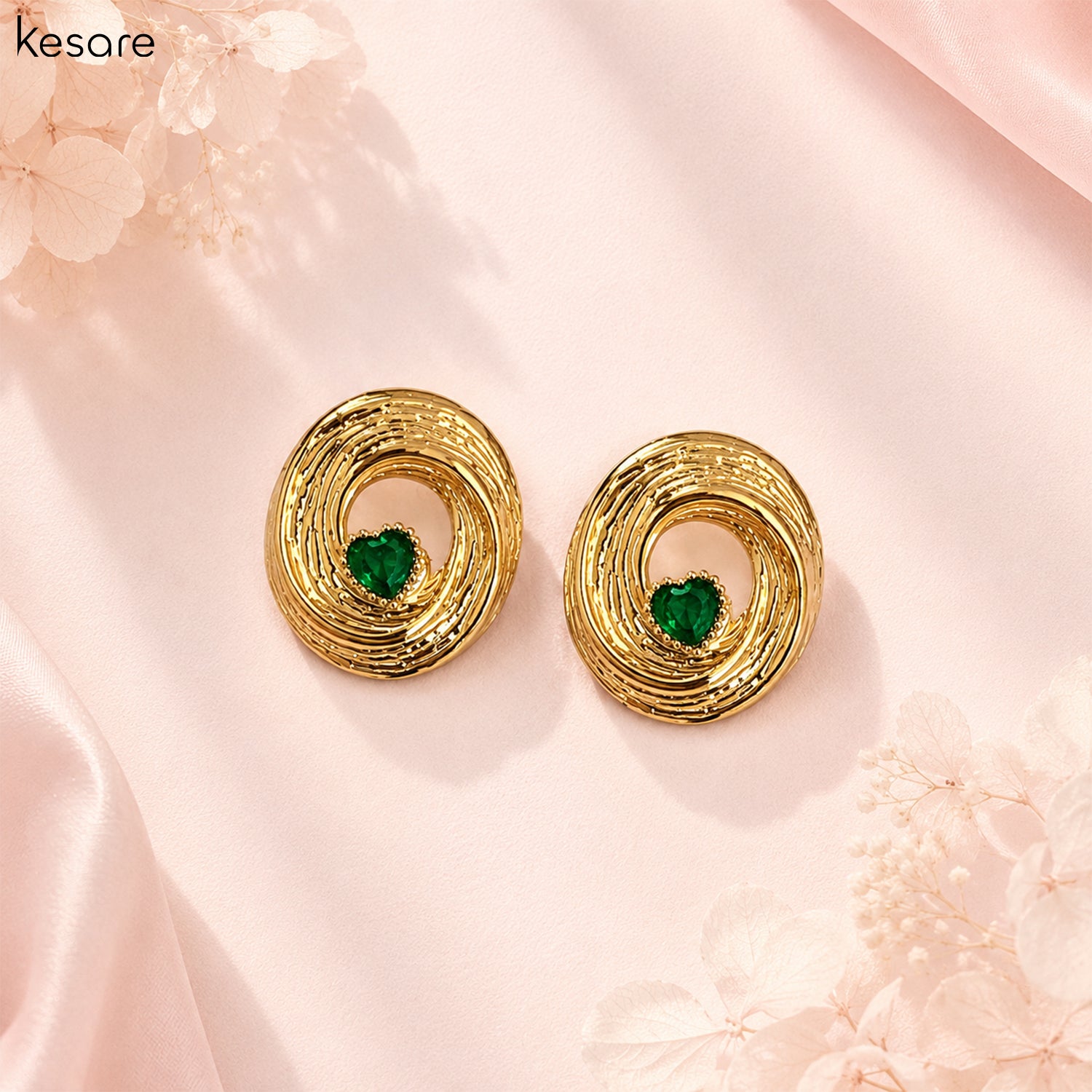 A pair of gold-toned spiral stud earrings with a small green heart-shaped stone in the center, displayed on a soft pink satin background with decorative floral elements.