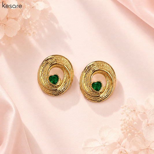 A pair of gold-toned spiral stud earrings with a small green heart-shaped stone in the center, displayed on a soft pink satin background with decorative floral elements.