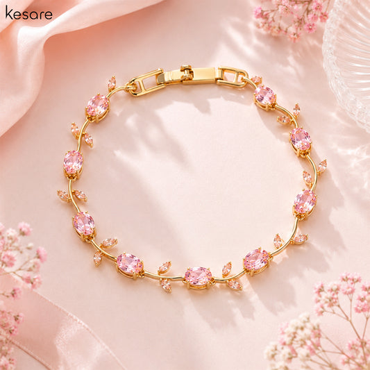 A gold-plated bracelet with pink rhinestones arranged in a floral vine pattern, displayed on a soft pink satin background with decorative flowers and ribbons.
