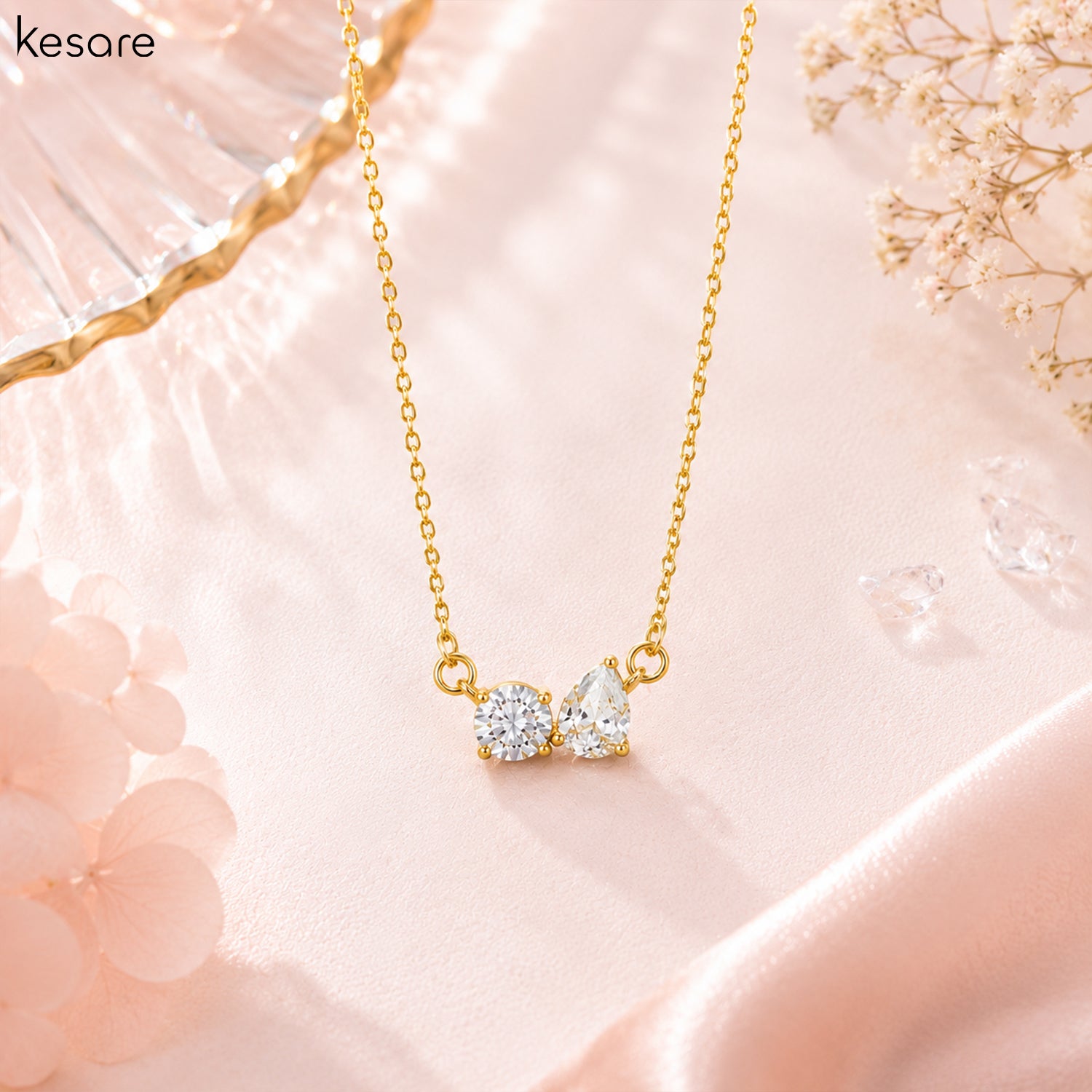 A gold necklace with a double gemstone pendant on a soft pink satin background. The pendant features two clear, sparkling stones in gold settings.