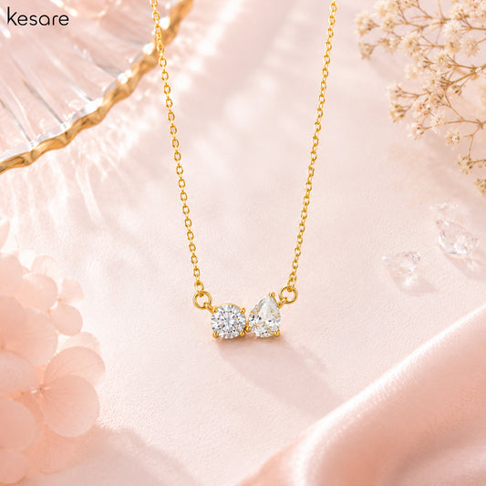 A gold necklace with a double gemstone pendant on a soft pink satin background. The pendant features two clear, sparkling stones in gold settings.