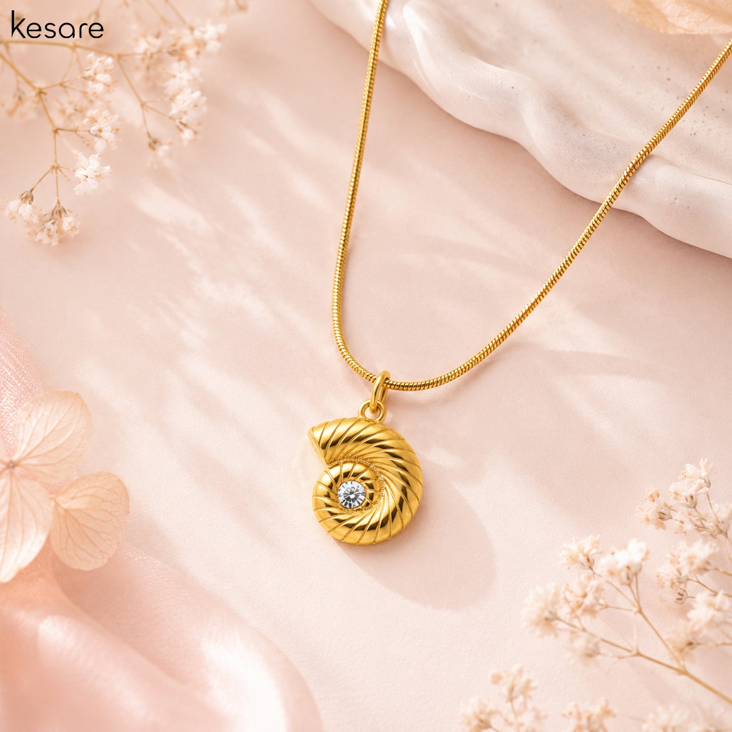 A gold necklace with a spiral shell-shaped pendant featuring a small clear stone in the center, displayed on a soft pink background with decorative dried flowers. The brand name 'Kesare' is visible in the top left corner.