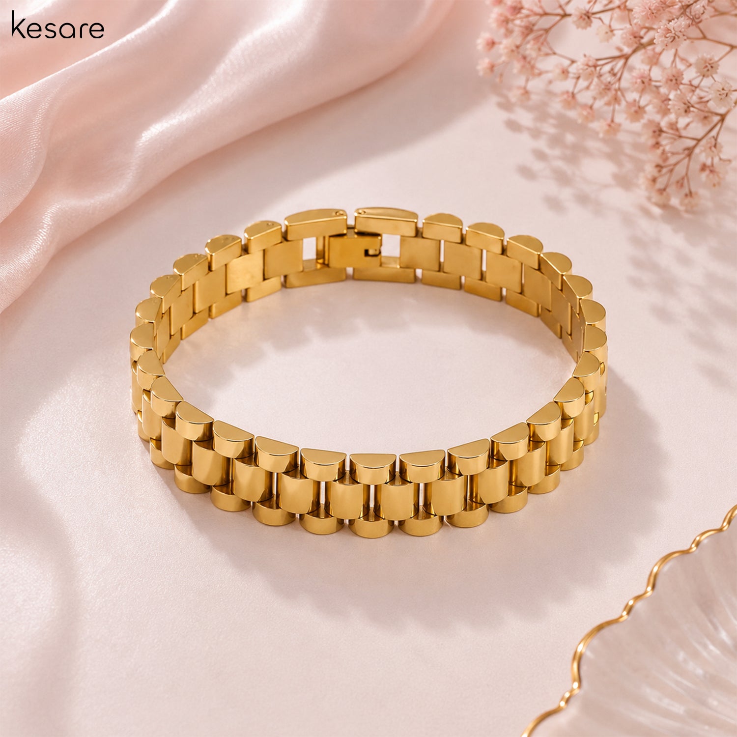 A gold-plated metal link bracelet with a polished finish, displayed on a soft beige fabric background with decorative pink satin and dried flowers. The brand name 'Kesare' is visible in the top left corner.