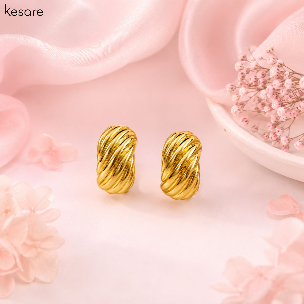 A pair of gold twisted rope stud earrings displayed on a soft pink background with decorative satin fabric, dried flowers, and a small dish.
