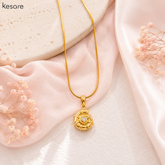 A gold pendant necklace with a floral rose design and a central clear stone, displayed on soft pink satin fabric with dried pink flowers. The brand name 'Kesare' is visible in the top left corner.