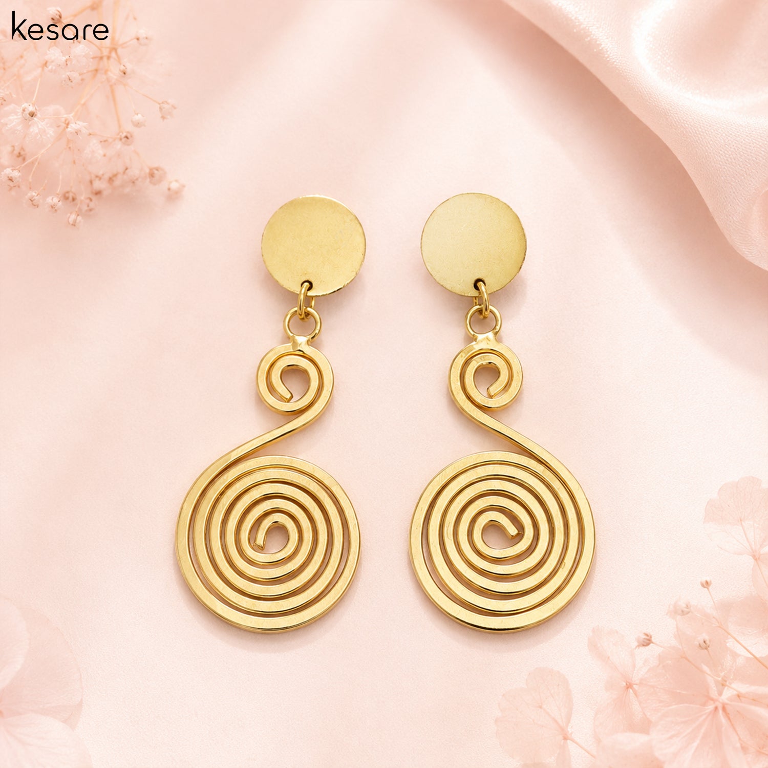 A pair of gold spiral drop earrings with flat circular studs, displayed on a soft pink satin background with decorative floral elements. The brand name 'Kesare' is visible in the top left corner.