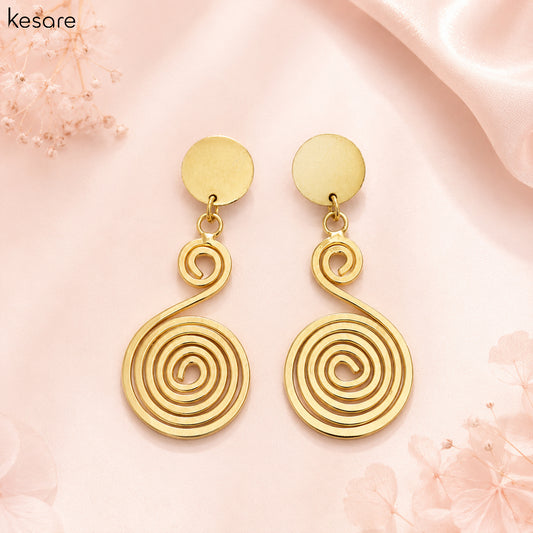 A pair of gold spiral drop earrings with flat circular studs, displayed on a soft pink satin background with decorative floral elements. The brand name 'Kesare' is visible in the top left corner.