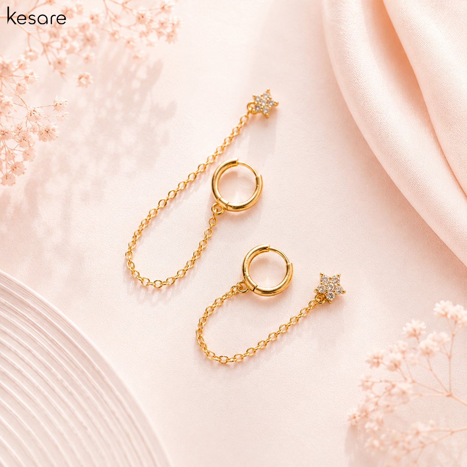 A pair of gold-toned drop earrings with circular ring charms and star-shaped drops, styled on a soft pink background with decorative dried flowers and satin fabric.