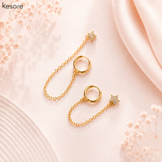 A pair of gold-toned drop earrings with circular ring charms and star-shaped drops, styled on a soft pink background with decorative dried flowers and satin fabric.