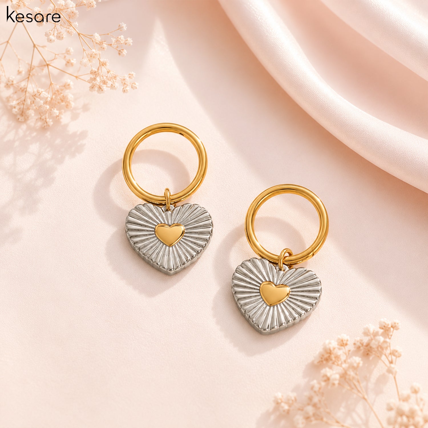 A pair of Kesare heart-shaped drop earrings with textured silver outer halves and a polished gold center heart, attached to gold rings, displayed on a soft pink background with dried flowers.