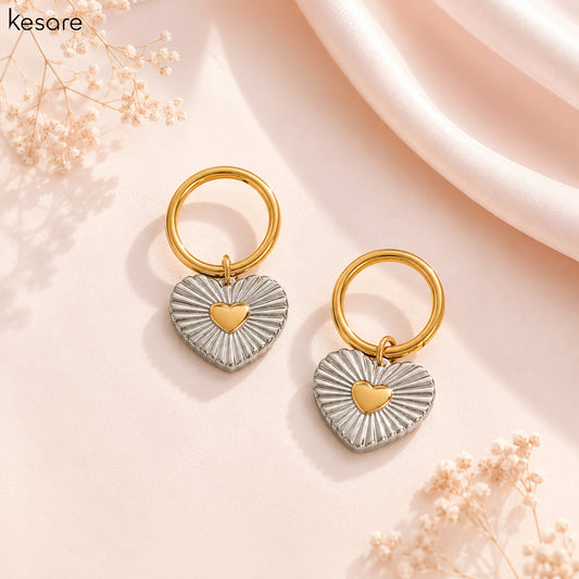 A pair of Kesare heart-shaped drop earrings with textured silver outer halves and a polished gold center heart, attached to gold rings, displayed on a soft pink background with dried flowers.